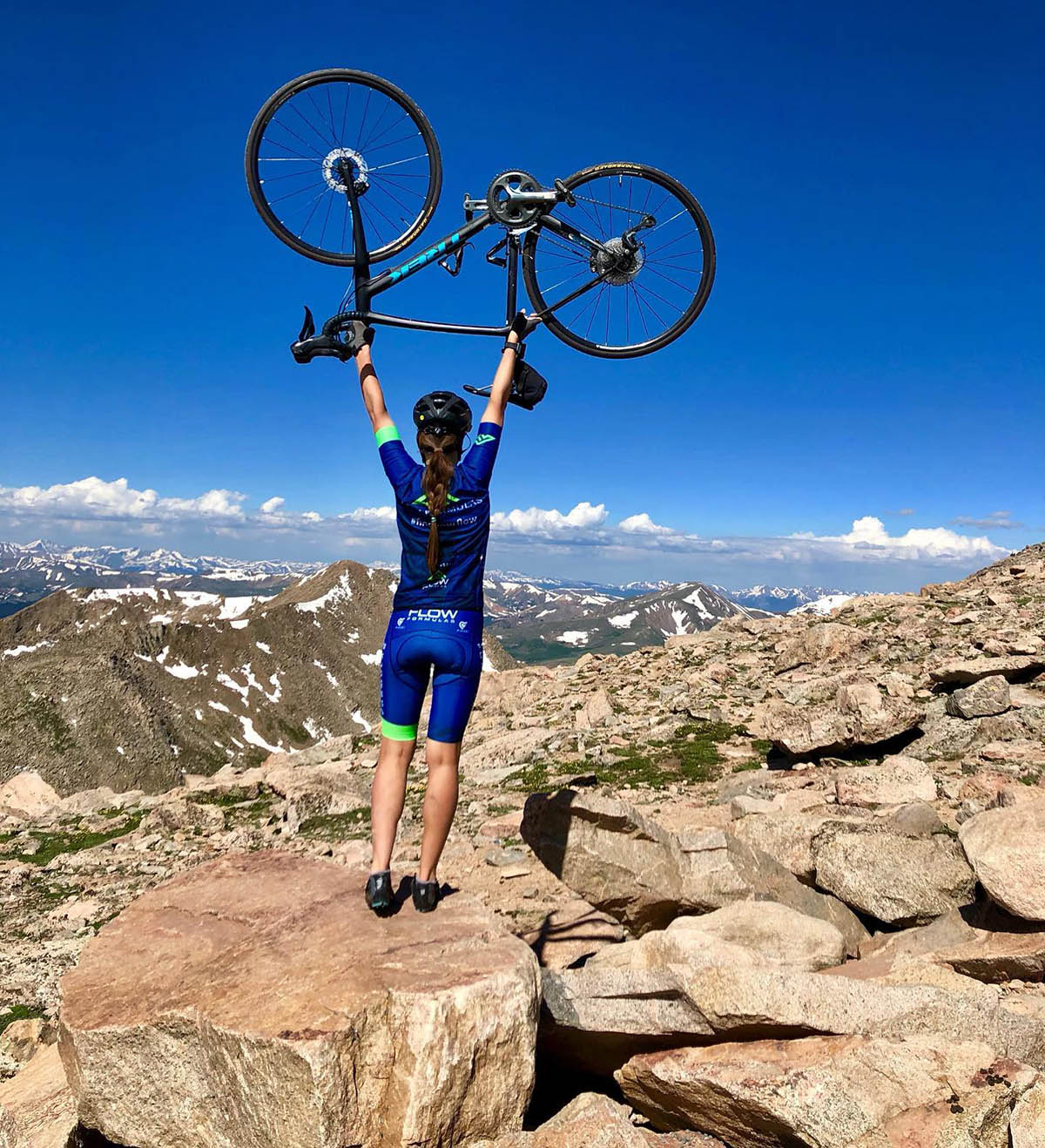 A biker stands majestically atop high-mountain rocks, holding her bike over her head as a sign of triumphant accomplishment.